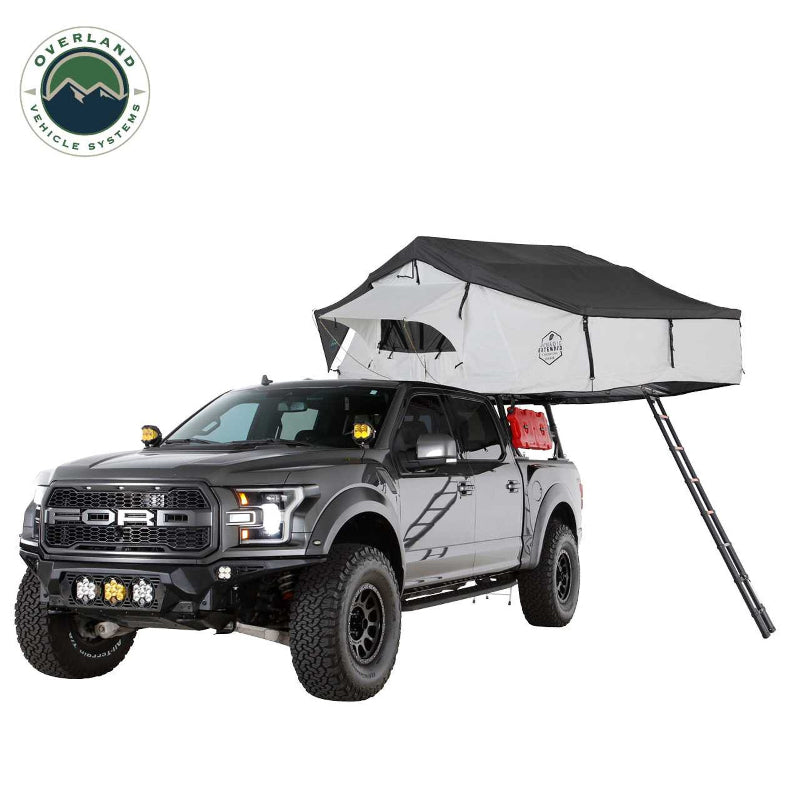 Image of a grey Ford pickup truck with a white and dark grey Nomadic Extended rooftop tent fully deployed on its roof, including an attached ladder extending to the ground, set against a white background