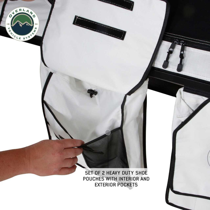 Image of a hand opening one of two white heavy-duty shoe pouches with interior and exterior pockets, hanging from a Nomadic rooftop tent, visible from a close-up, angled view