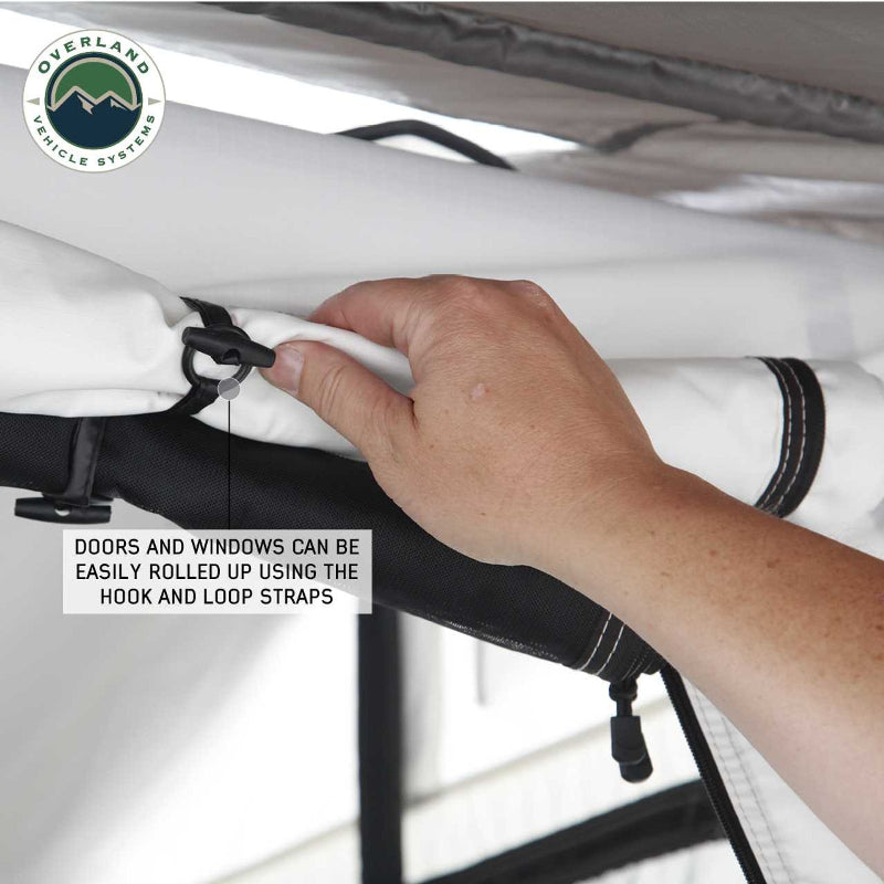 Image of a hand rolling up a white tent door or window, secured with a black hook and loop strap on a Nomadic rooftop tent, visible from a close-up, angled view