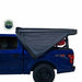 A gray Nomadic Awning is partially deployed on the side of a blue pickup truck. The awning is shown from an angled side and rear view, with the Overland Vehicle Systems logo visible on the side of the truck bed