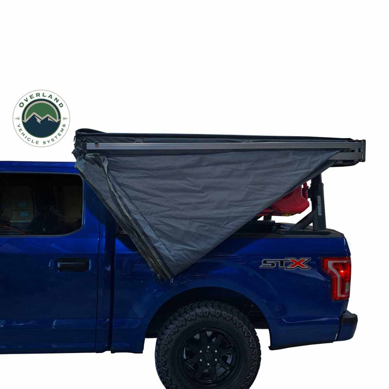 A gray Nomadic Awning is partially deployed on the side of a blue pickup truck. The awning is shown from an angled side and rear view, with the Overland Vehicle Systems logo visible on the side of the truck bed