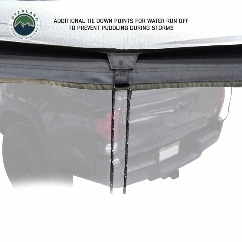 Image of the Overland Vehicle Systems Nomadic Awning with a close-up view of the black webbing tie-down point and a length of paracord hanging from it. The description "ADDITIONAL TIE DOWN POINTS FOR WATER RUN OFF TO PREVENT PUDDLING DURING STORMS" is visible above the tie-down point, highlighting a key feature of the awning