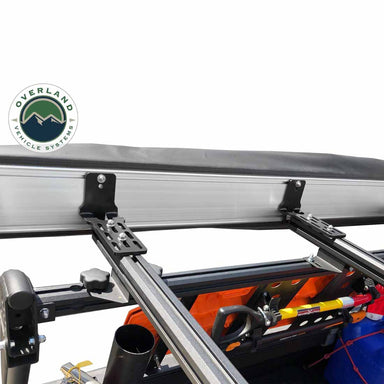 Image of the OVS Nomadic 180° Awning mounting brackets, including black L-shaped and slotted flat brackets, attached to a vehicle's roof rack with other gear visible
