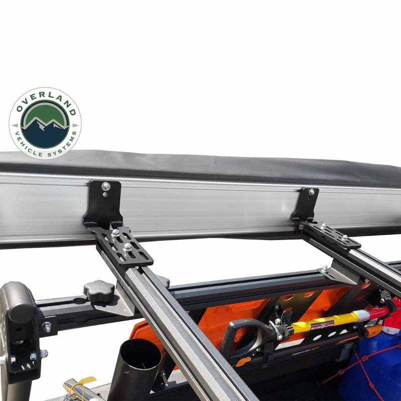 Image of the OVS Nomadic 180° Awning mounting brackets, including black L-shaped and slotted flat brackets, attached to a vehicle's roof rack with other gear visible