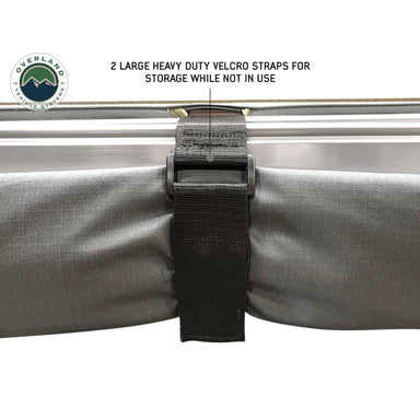 Image of a gray awning fabric rolled up and secured by a black heavy-duty velcro strap. The text "2 LARGE HEAVY DUTY VELCRO STRAPS FOR STORAGE WHILE NOT IN USE" is visible, along with the "OVERLAND VEHICLE SYSTEMS" logo