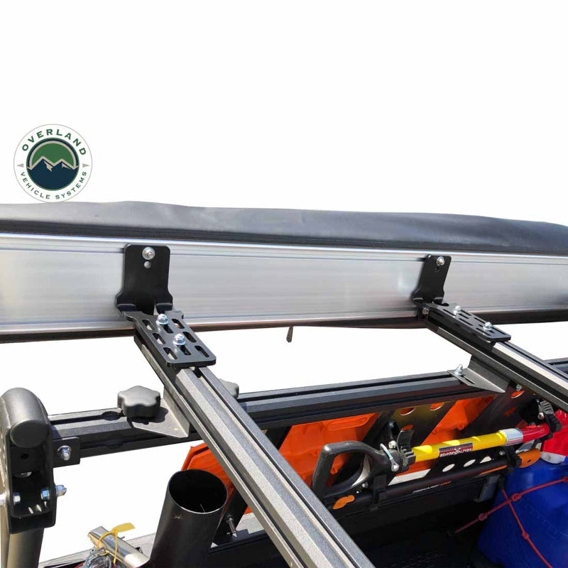 Image of a close-up of the mounting bracket for a rooftop tent or awning, attached to a roof rack. An orange traction board, a shovel, and other off-road accessories are visible underneath