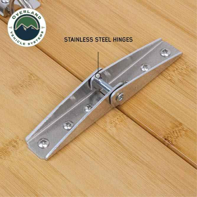 Image of OVS Kick It bi-fold camp table hinge detail showing stainless steel folding hinge mounted on bamboo wood tabletop, close-up angled view on a white background.