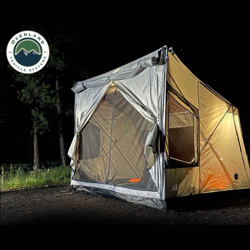 A lit-up OVS ground tent is set up at night in a wooded area. The interior lights illuminate the tent, showcasing the mesh windows and the gray fabric