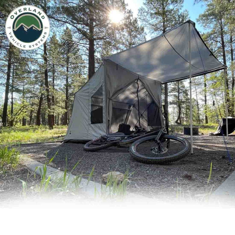 An angled front view of the OVS ground tent shows it deployed in a forest campsite with an awning providing shade. A mountain bike is leaning against the tent, set on a gravel pad with trees in the background and sunlight shining down