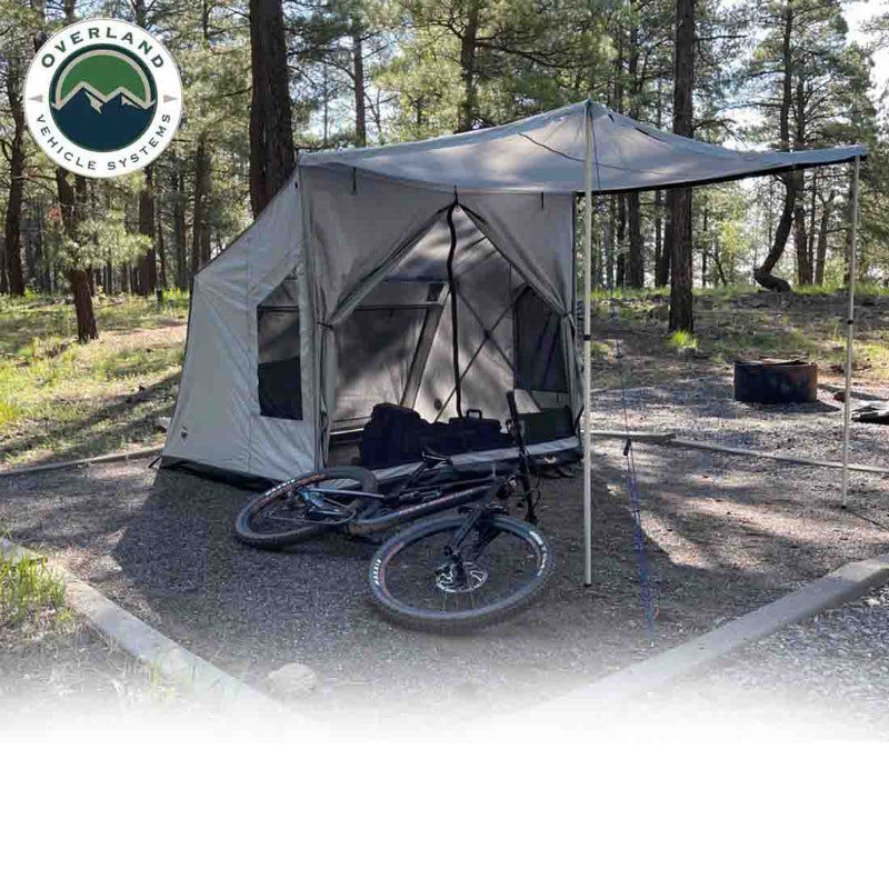 An angled front view of the OVS ground tent shows it deployed in a forest campsite with an awning providing shade. A mountain bike and gear are seen inside the front door, set on a gravel pad with a fire pit nearby and trees in the background