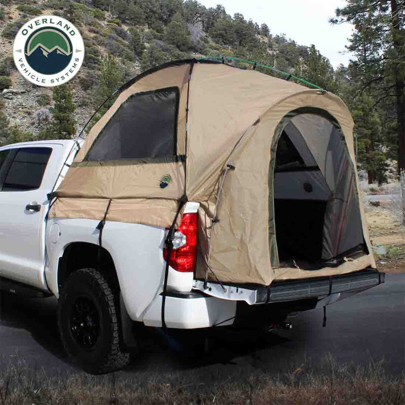 Image of a pickup truck bed tent installed on a white truck, tan fabric with mesh windows and open rear entry, angled rear and side view in an outdoor mountain setting.
