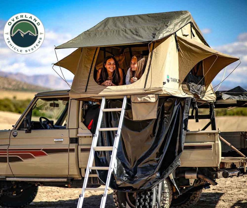 Image of two smiling women inside a rooftop tent mounted on a tan overland vehicle, with a ladder providing access, set against a desert landscape.