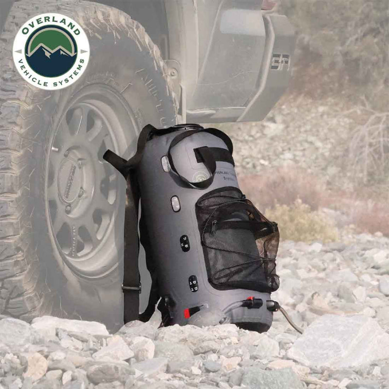 Image of Overland Vehicle Systems portable camp shower bag standing upright next to an off-road vehicle tire, gray waterproof design with mesh pocket and straps shown in outdoor rocky terrain setting