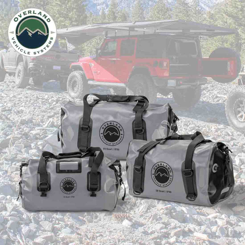 Image of three gray Overland Vehicle Systems IPX6 waterproof duffel bags (69, 42, and 58 Quart) on a rocky terrain background with an overland vehicle and tent in the soft-focus distance.