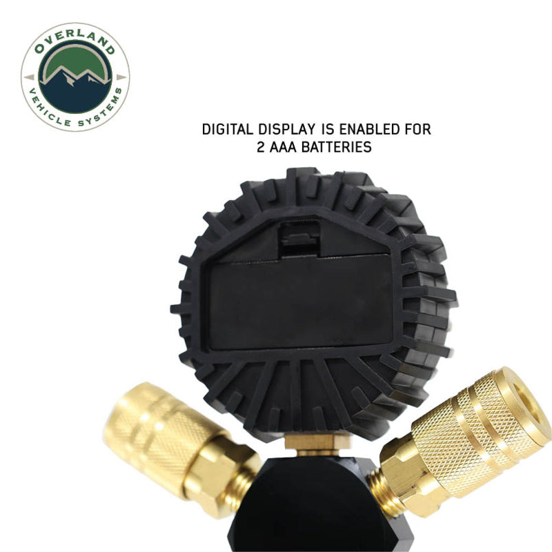 An image showing the back of the Overland Vehicle Systems (OVS) D.T.D. digital tire deflator's main unit, with a rectangular battery compartment cover visible. The text "DIGITAL DISPLAY IS ENABLED FOR 2 AAA BATTERIES" is displayed above the unit, with brass quick connect fittings visible at the bottom. The OVS logo is in the top left