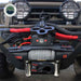 Overland Vehicle Systems (OVS) SCAR Series Winch installed on the front bumper of a gray vehicle. The winch is black with orange accents, featuring synthetic rope, a hook, and a red pulley system, with two large off-road lights mounted above it