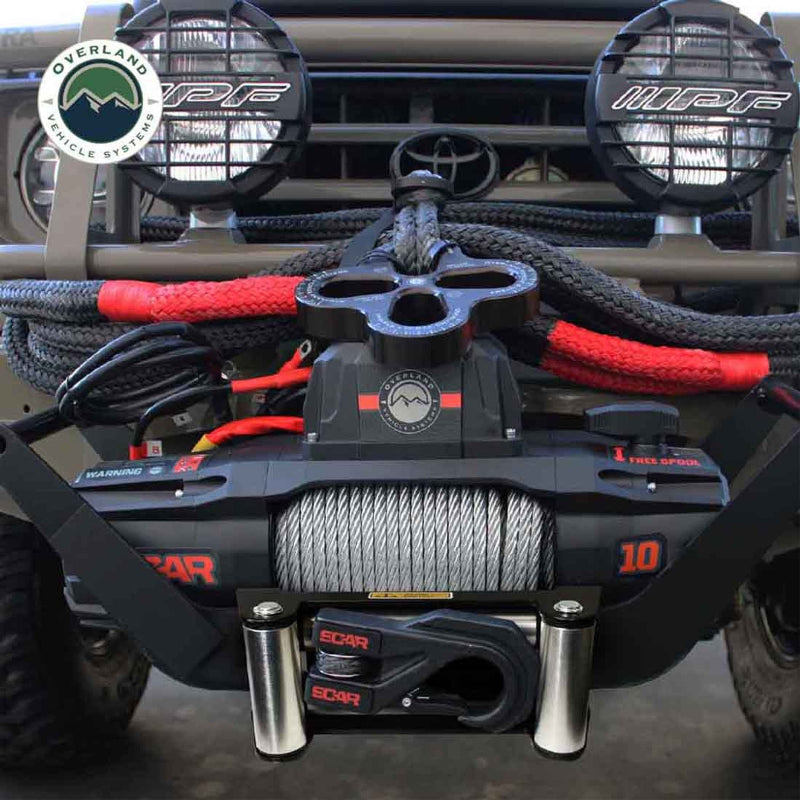Overland Vehicle Systems (OVS) SCAR Series Winch installed on the front bumper of a gray vehicle. The winch is black with orange accents, featuring synthetic rope, a hook, and a red pulley system, with two large off-road lights mounted above it