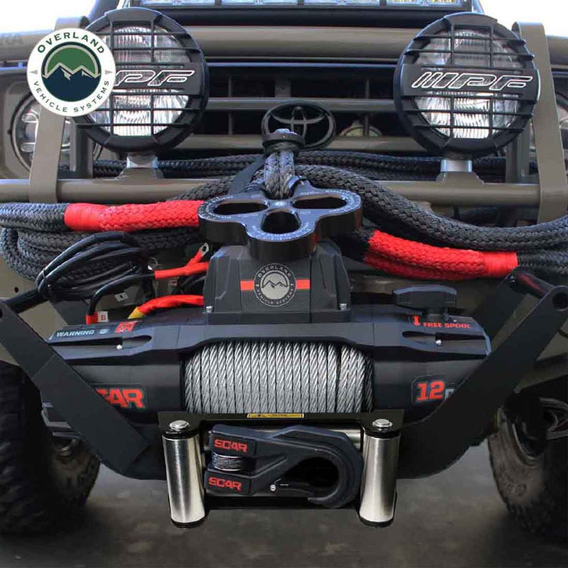 Image of an OVS SCAR 12 Steel Cable Winch installed on the front of a dark green off-road vehicle, with a close-up, front view. The winch has a steel cable spooled, a black fairlead with a hook, and a tree saver strap wrapped around the winch line. Two round off-road lights are visible above the winch