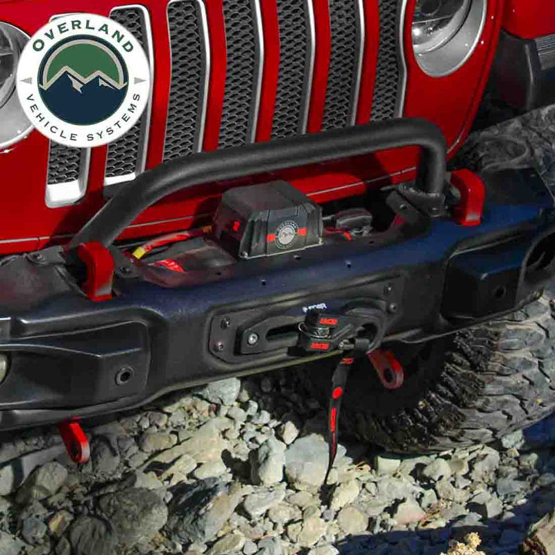 Image of an Overland Vehicle Systems (OVS) SCAR winch installed on the front bumper of a red Jeep Wrangler, with a hook connected to a winch line. The vehicle is on a rocky, off-road surface