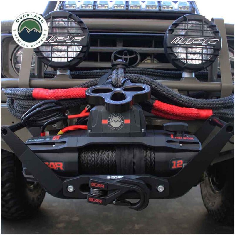 Image of the black Overland Vehicle Systems (OVS) SCAR winch installed on the front bumper of a truck, featuring a red '12p' and a shackle block for heavy-duty pulling. The truck has large, round spotlights and is ready for off-road use