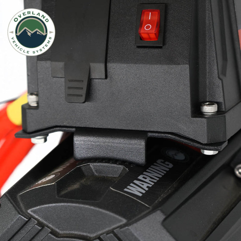 Image of a close-up on the black Overland Vehicle Systems (OVS) SCAR winch housing, highlighting the red power switch and a 'WARNING' label. The image shows the detail of the solenoid and its mounting bracket