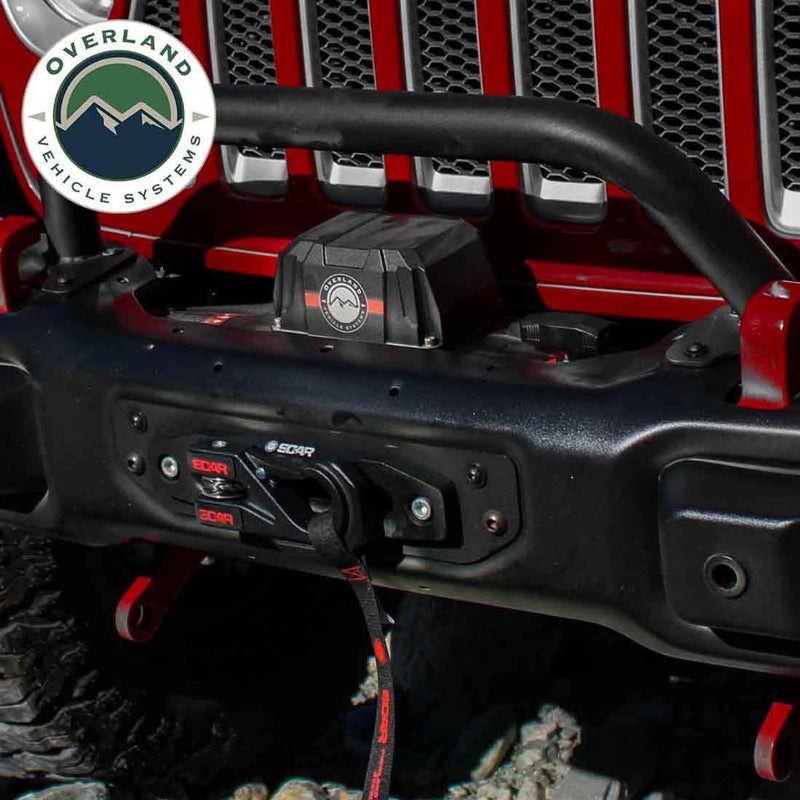 Image of an Overland Vehicle Systems (OVS) SCAR winch installed on a red Jeep Wrangler's front bumper, with the hook and strap connected to the fairlead. The winch is black with red accents and the Jeep is on a rocky, off-road surface