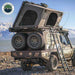 Image of an overland vehicle, possibly a Toyota Land Cruiser, with a dark grey hardshell rooftop tent fully deployed on its bed, featuring two large spare tires mounted at the rear and a ladder extended to the side. The vehicle is parked on a rocky shore with a body of water and mountains in the background, with the "Overland Vehicle Systems" logo in the top left