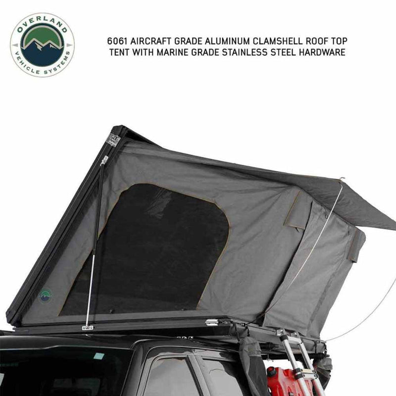 Image of an Overland Vehicle Systems 6061 Aircraft Grade Aluminum Clamshell Roof Top Tent, partially deployed with a dark grey fabric tent body and an extended awning, shown on a white background. The tent is mounted on a vehicle and highlights marine-grade stainless steel hardware
