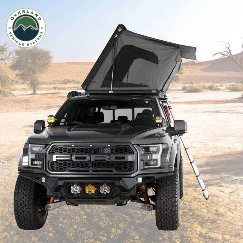 Image of a black Ford pickup truck with a dark grey hardshell rooftop tent partially deployed on its roof, positioned in a desert landscape with a ladder extended to the side. The truck features aftermarket off-road lights and a rugged front bumper, with the "Overland Vehicle Systems" logo in the top left