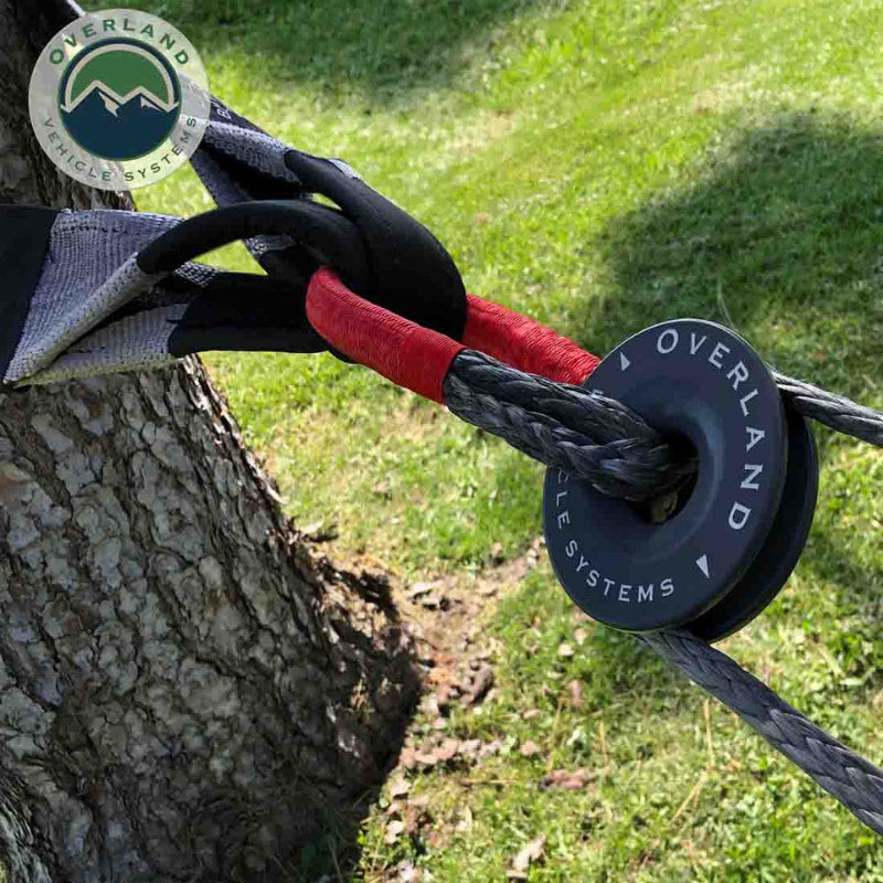 Image of Overland Vehicle Systems soft shackle in use securing a recovery strap to a tree, gray braided synthetic rope with black steel collar and red protective sleeve, outdoor angled close-up view.