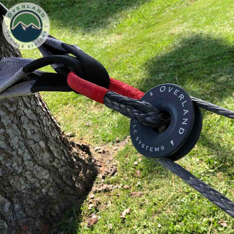Image of a soft shackle, with red protective sleeving, connected to a tree saver strap wrapped around a tree trunk, and threaded through a black recovery ring, with grass in the background and the "Overland Vehicle Systems" logo.