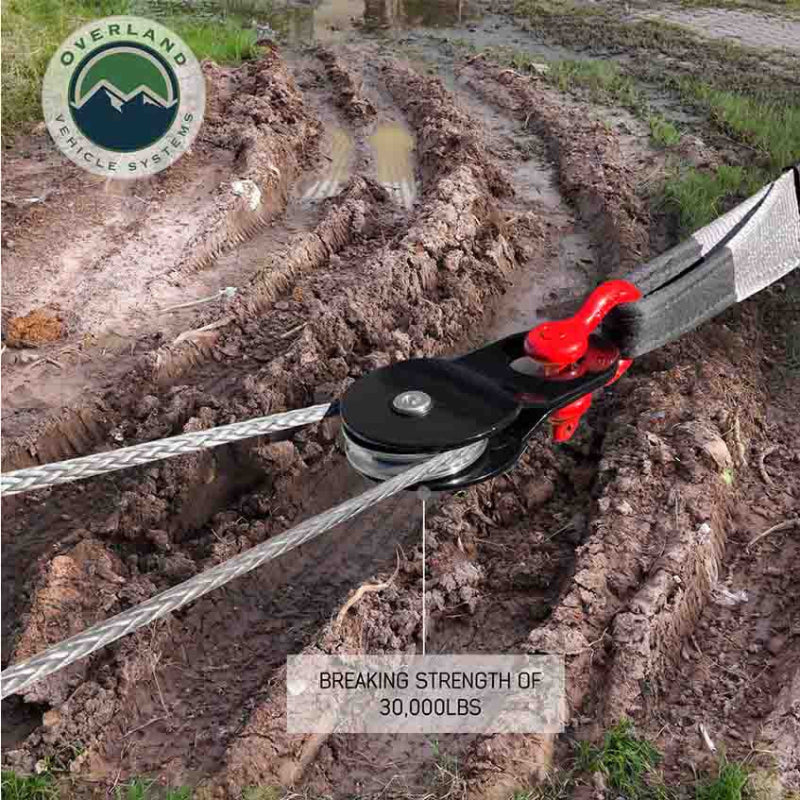 Image of Overland Vehicle Systems standard snatch block in use during vehicle recovery, black pulley with red shackle guiding winch cable through muddy terrain, angled close-up outdoor view.