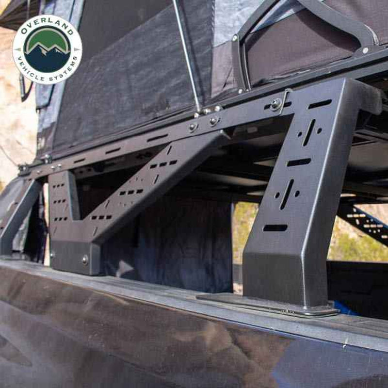Image of a close-up view of the heavy-duty support legs of a mid-height rack for a pickup truck, showcasing its robust black frame, positioned to securely hold a roof top tent, set against a rugged desert backdrop