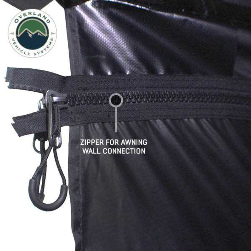 Image showcasing a black Overland Vehicle Systems 270-degree awning partially deployed from the side of a dark grey pickup truck, illustrating how the awning is stored and deployed. The awning fabric is folded and secured against the truck, indicating a travel position