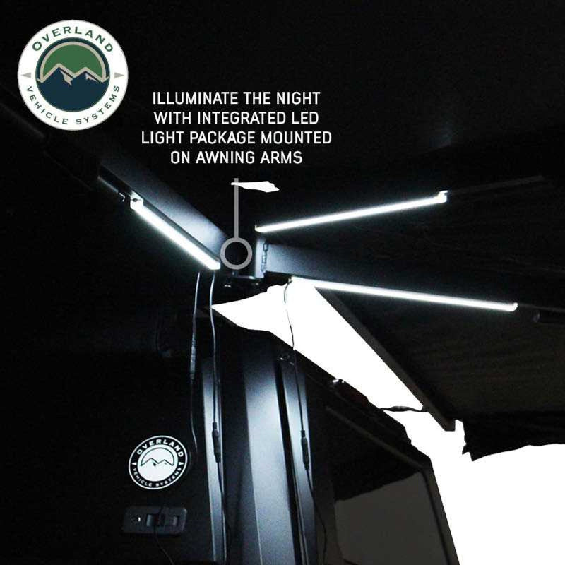Image displaying the underside of a black Overland Vehicle Systems awning, illuminated by an integrated LED light package mounted on the awning arms to light up the night. The "Overland Vehicle Systems" logo is in the top left corner