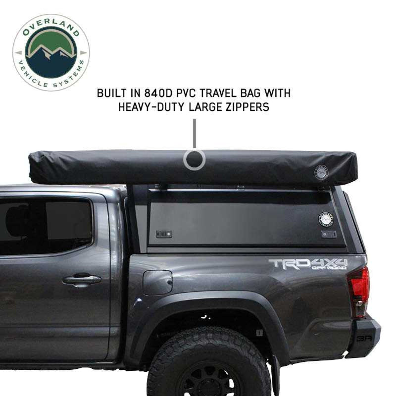 Image showcasing a black Overland Vehicle Systems 270-degree awning in a built-in 840D PVC travel bag with heavy-duty large zippers, mounted on top of a pickup truck bed cap. The vehicle is a dark grey Toyota Tacoma with "TRD 4X4 OFF ROAD" written on the side