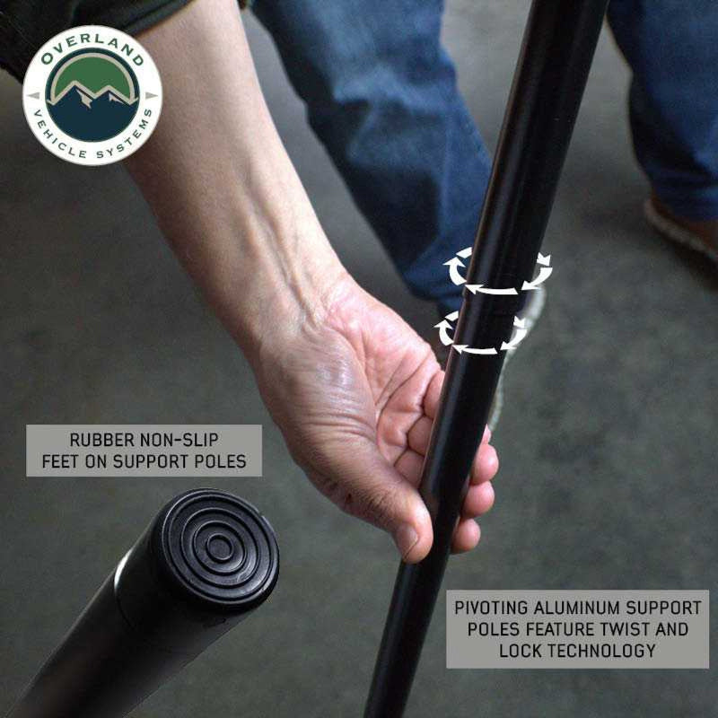 Image showcasing the underside of a black Overland Vehicle Systems awning, highlighting its pivoting aluminum support poles with twist and lock technology and rubber non-slip feet. The "Overland Vehicle Systems" logo is in the top left corner