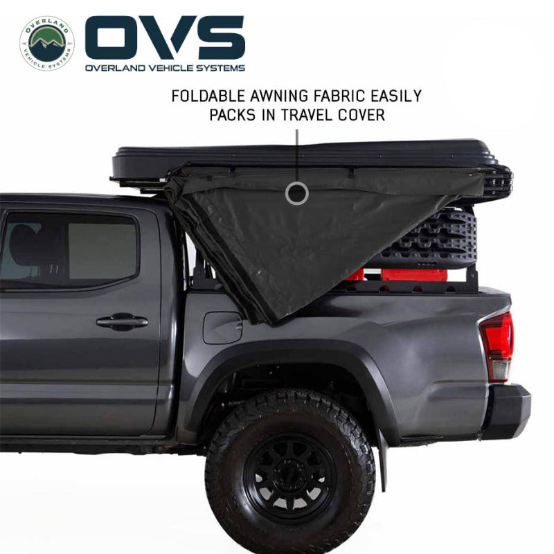 Image of an OVS XD Nomadic 270 LTE awning partially folded and packing into its travel cover on a pickup truck, black fabric shown mid-collapse in side view on a white background.
