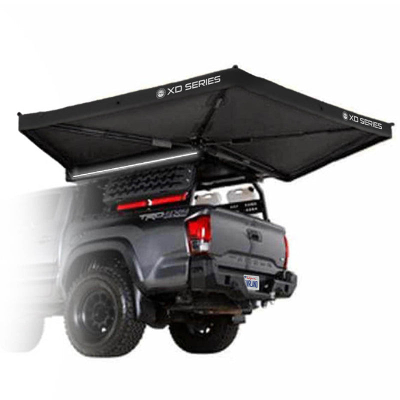 Image of an OVS XD Nomadic 270 LTE vehicle-mounted awning fully deployed from the rear of a pickup truck, black fabric with integrated LED lighting, shown in angled rear and side view on a white background.