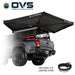 Image of an OVS XD Nomadic 270 LTE awning deployed from the rear of a pickup truck, black fabric with included LED lighting strip illuminated underneath, shown in angled rear and side view on a white background.