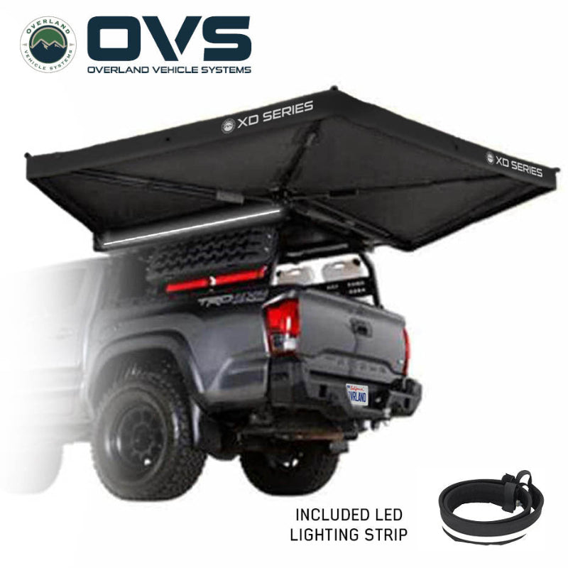 Image of an OVS XD Nomadic 270 LTE awning deployed from the rear of a pickup truck, black fabric with included LED lighting strip illuminated underneath, shown in angled rear and side view on a white background.