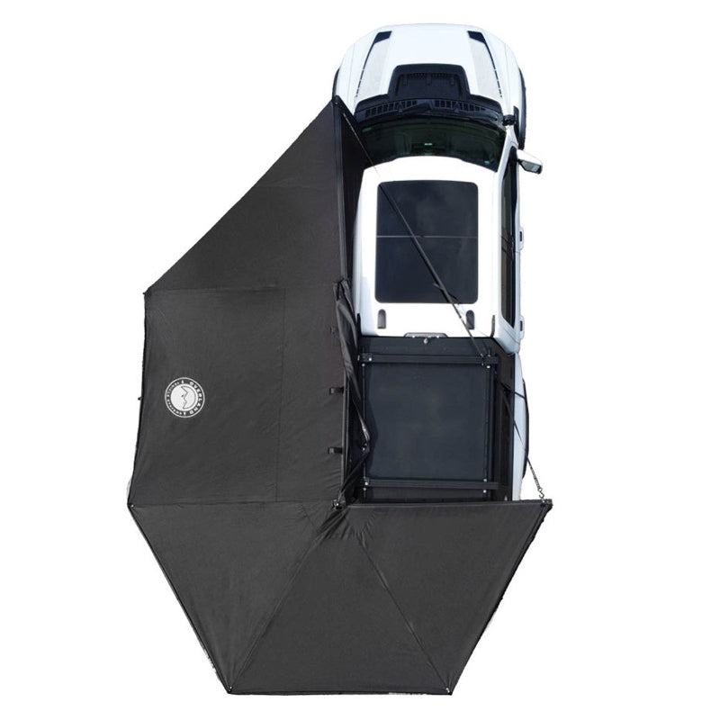 Top-down view of a white SUV with an OVS Nomadic Awning partially deployed. The black awning is attached to the side of the vehicle, extending outwards and downwards to create a covered space