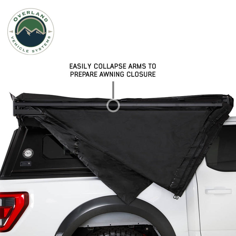 Side view of a white pickup truck with the OVS Nomadic Awning in the process of being stowed away. The black awning fabric is being collapsed, with an annotation explaining that the arms are being folded to prepare for closure