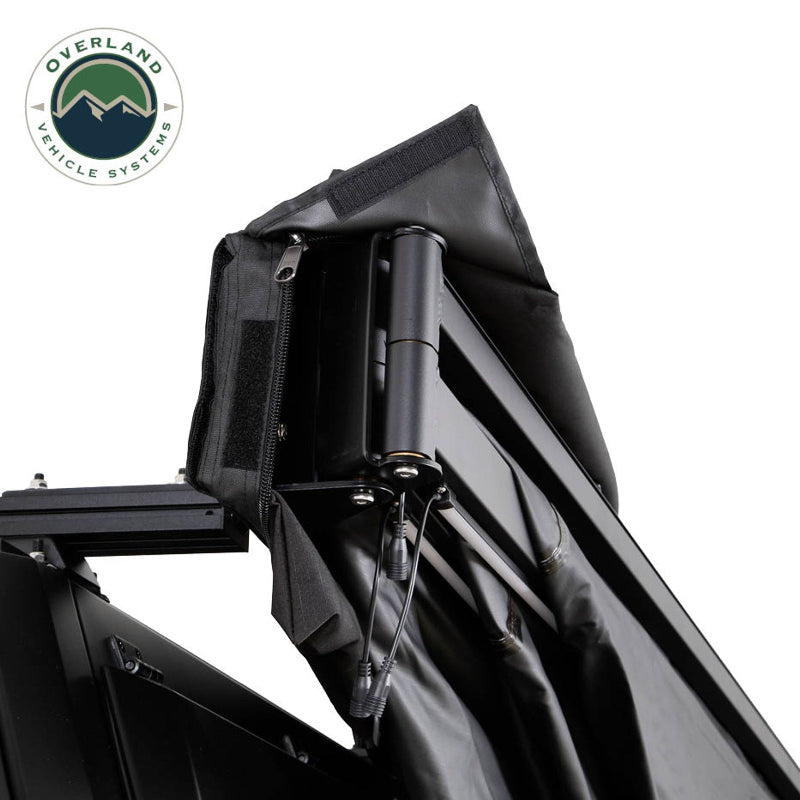 Close-up, angled view of the OVS Nomadic Awning, showing the internal hardware and LED wiring. The black awning fabric is partially open, revealing the black support arms and the integrated LED lighting system's cables