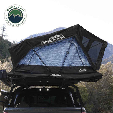 Image of an Overland Vehicle Systems XD Series Sherpa Roof Top Tent, fully deployed on the back of a pickup truck in a mountainous outdoor setting. The tent is black with a light blue patterned interior visible through the mesh windows, and the "SHERPA" logo is prominently displayed on the front