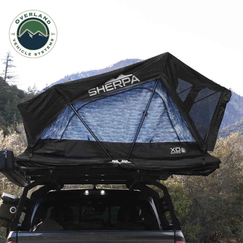 Image of an Overland Vehicle Systems XD Series Sherpa Roof Top Tent, fully deployed on the back of a pickup truck in a mountainous outdoor setting. The tent is black with a light blue patterned interior visible through the mesh windows, and the "SHERPA" logo is prominently displayed on the front