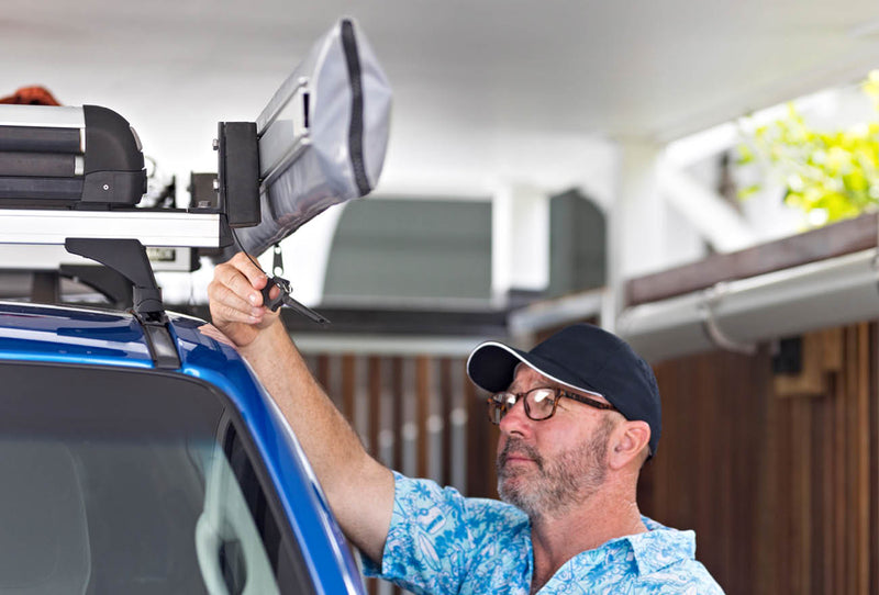Image of a bearded man in a blue floral shirt, glasses, and a black baseball cap, attaching a grey and black awning to the roof rack of a blue vehicle.