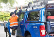 Image of a tradesman in a high-visibility vest and cap, wearing a tool belt, reaching up to secure a long, light-colored tube onto the roof rack of a blue 4x4 pickup truck with a canopy, in an outdoor setting.