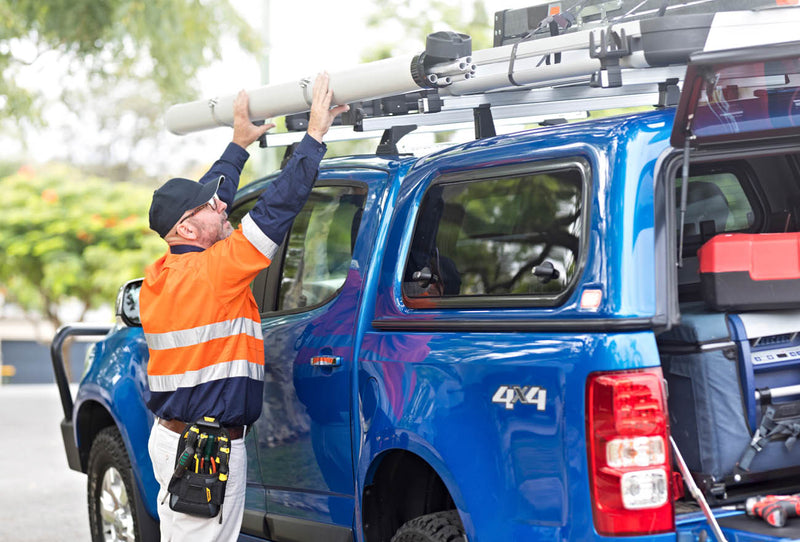 Image of a tradesman in a high-visibility vest and cap, wearing a tool belt, reaching up to secure a long, light-colored tube onto the roof rack of a blue 4x4 pickup truck with a canopy, in an outdoor setting.