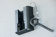 Image of a black Rack 'N' Trax awning bracket mounted on a light gray textured surface, with a key inserted and a security cable attached.